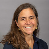 Anne Delano Steinert, a white woman with long brown hair, wears a blue shirt, earrings and necklace